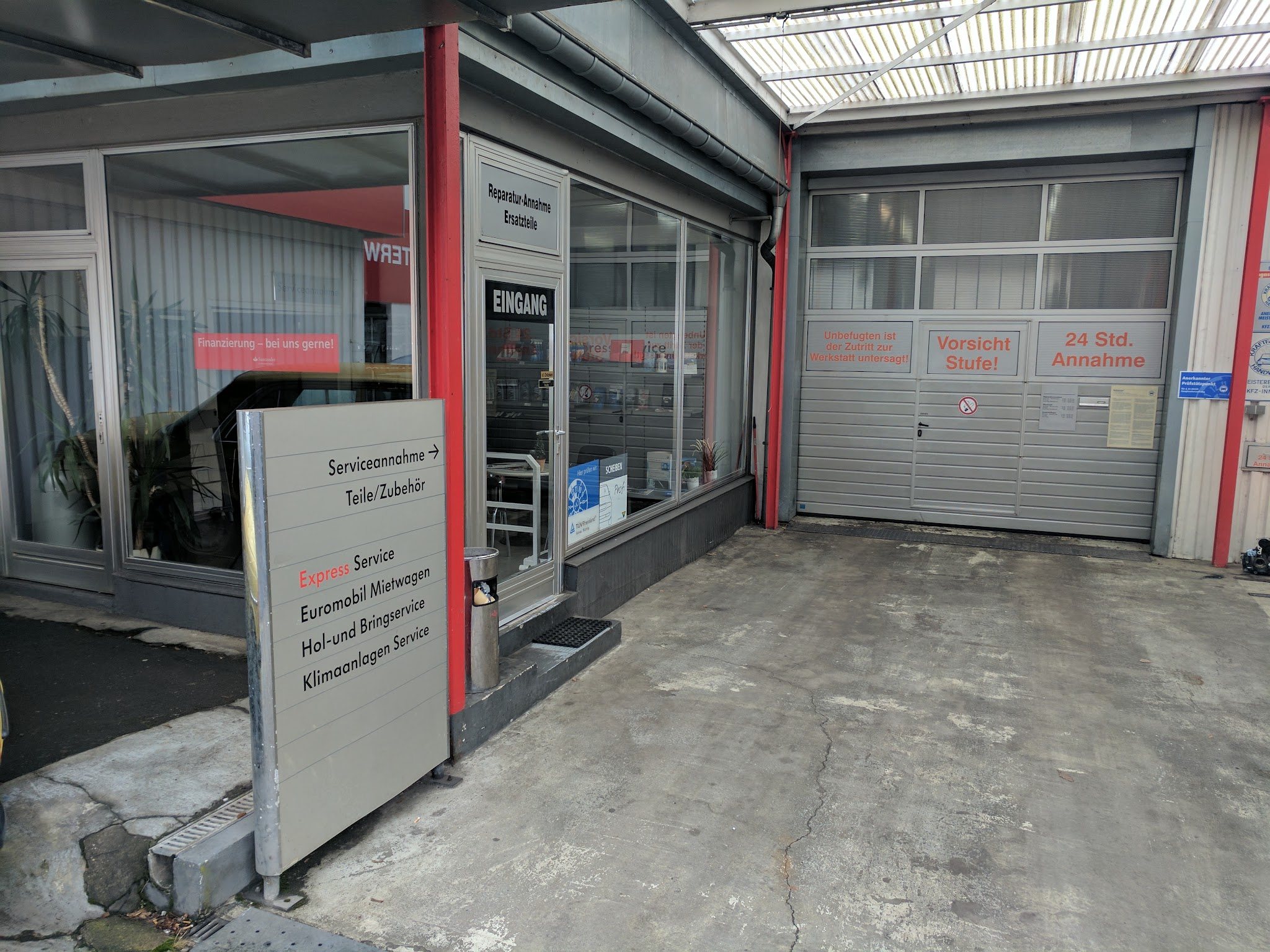 Eingang der Werkstatt mit Leistungsschild, Rolltor mit der Aufschrift 24 Std. Annahme und TÜV-Rheinland-Aufkleber